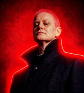 Artistic portrait of Tammy Eddy with bright pink hair and a dark coat, outlined by a glowing red neon light against a smokey red background.