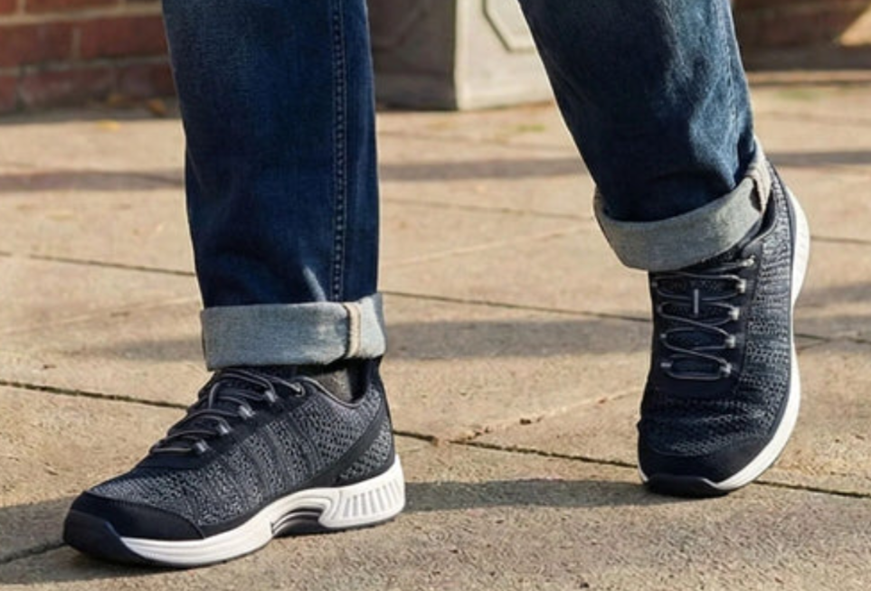 Person wearing rolled-hem jeans and black knit sneakers walking on a paved sidewalk.