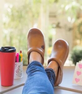 Person wearing tan leather nursing clogs or stable medical shoes resting their feet on a desk near a coffee mug.