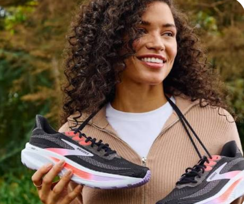 Woman with curly hair smiling while holding a pair of black and pink running shoes outdoors